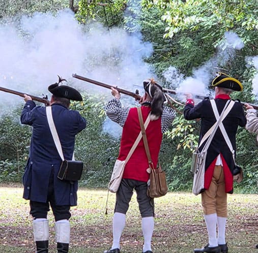 Reenactors in Revolutionary War uniforms firing muskets in a historical reenactment.
