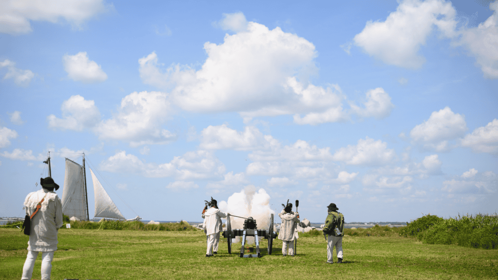 Historical reenactors in colonial attire with a cannon and sailboat in a grassy field.
