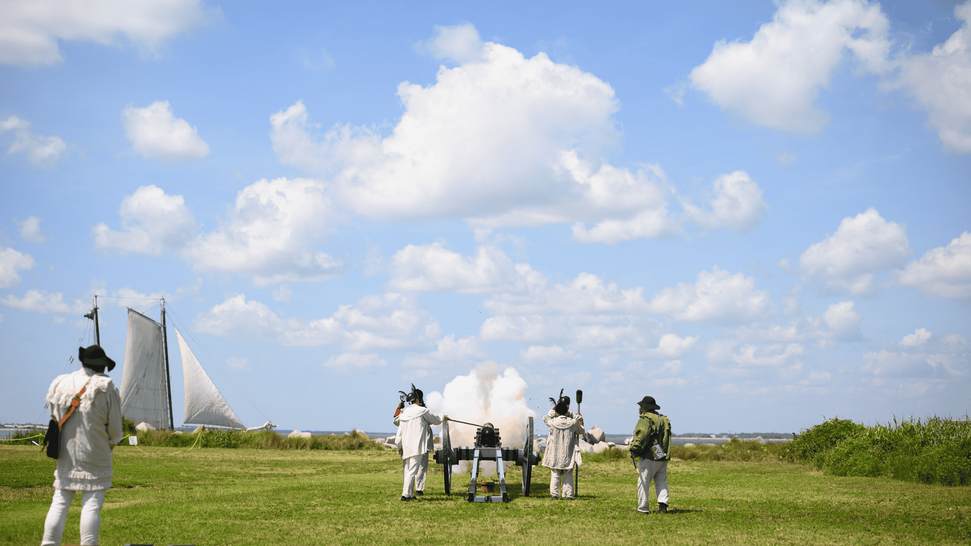 Untitled design (7) Historical reenactors in colonial attire with a cannon and sailboat in a grassy field.