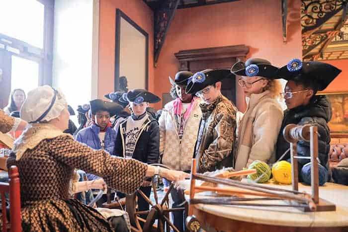 Students in tricorn hats watch a historical textile demonstration.